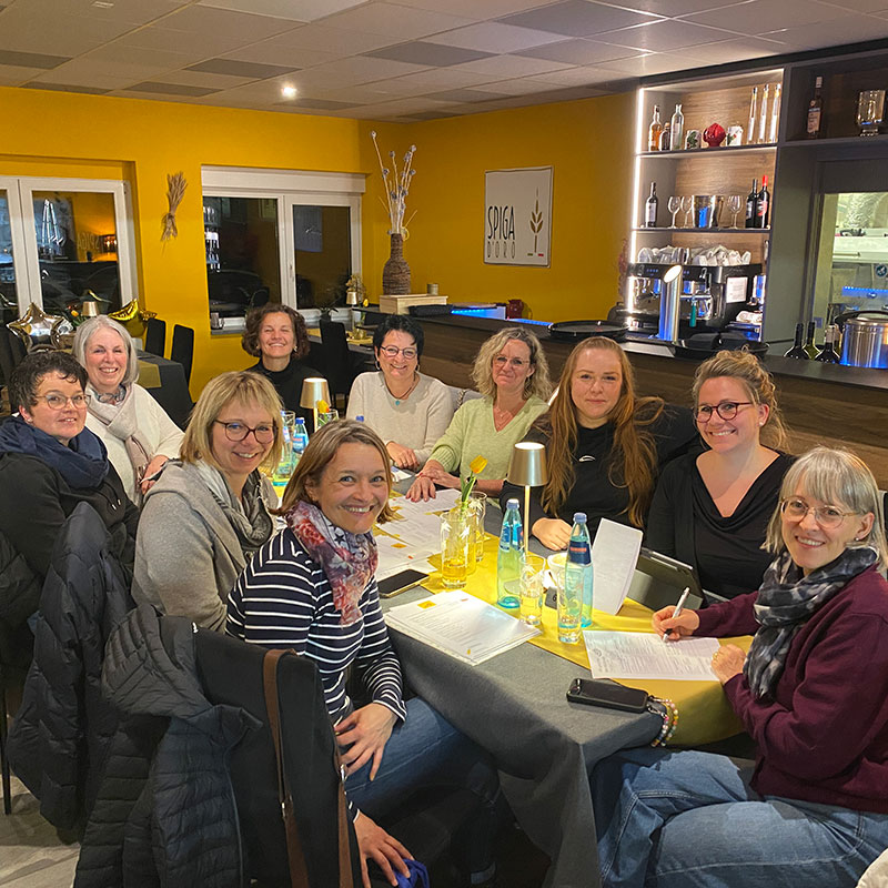 Gruppe von zehn Frauen sitzt gemeinsam an einem Tisch in einem gemütlichen Restaurant mit gelber Wand und modernem Barbereich im Hintergrund. Auf dem Tisch stehen Wasserflaschen, Gläser und Speisekarten. Alle lächeln in die Kamera und wirken entspannt und fröhlich.