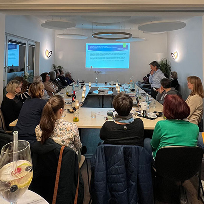 Personengruppe sitzt in einem modern eingerichteten Konferenzraum um einen großen, U-förmigen Tisch. Auf einer weißen Wand wird eine Präsentation projiziert. Die Teilnehmer*innen hören aufmerksam einem Vortrag zu. Auf dem Tisch stehen Getränke, Teelichter und Notizmaterialien.