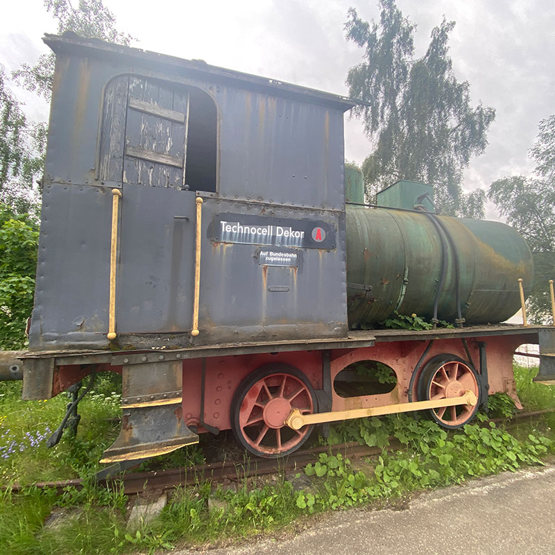 Eine alte, rostige Lokomotive steht auf einem kurzen Gleisstück im Freien, umgeben von Gras und Unkraut. Auf der Seite des Führerhauses ist ein Schild mit der Aufschrift ‚Technocell Dekor‘ sowie dem Hinweis ‚Auf Bundesbahn zugelassen‘ angebracht. Die Lok hat einen zylindrischen Tank hinter dem Führerhaus und ist sichtbar verwittert, mit abgeblätterter Farbe und Rostspuren.