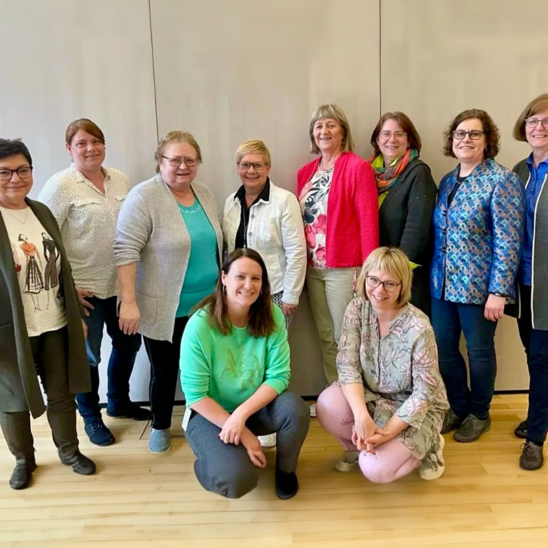 Gruppenfoto von zehn Frauen vor einer hellen Holz- oder Paneelwand. Acht Frauen stehen in einer Reihe, zwei sitzen davor auf dem Boden. Alle lachen oder lächeln in die Kamera. Die Kleidung ist bunt und lässig, was auf ein freundliches, informelles Treffen hinweist.
