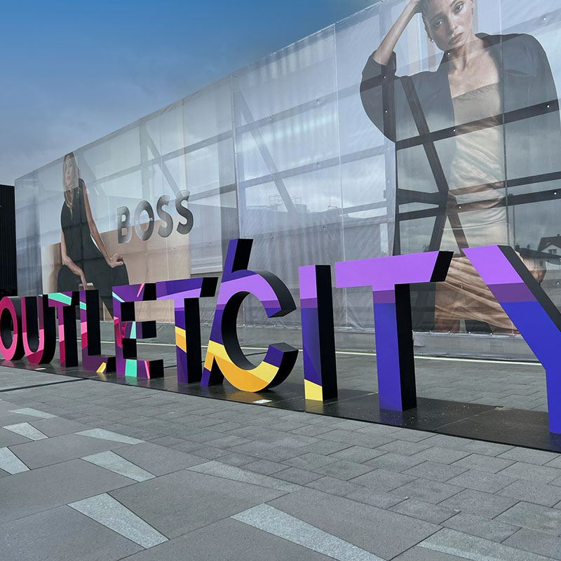 Große, bunte 3D-Buchstaben formen das Wort ‚OUTLETCITY‘ vor einer transparenten Fassade mit übergroßen Modeplakaten, auf denen Models in stilvoller Kleidung zu sehen sind. Im Hintergrund ist Werbung der Marke ‚BOSS‘ sichtbar. Der Boden besteht aus grauen Steinplatten unter einem teils bewölkten Himmel.