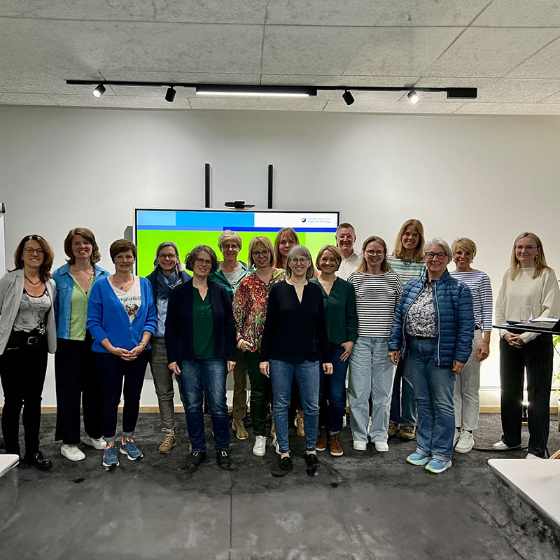 Gruppenfoto von 17 Personen (überwiegend Frauen) in einem modern eingerichteten Raum mit weißer Wand und großem Bildschirm im Hintergrund. Alle stehen in zwei Reihen, lächeln in die Kamera und tragen legere Kleidung. Der Bildschirm zeigt eine Präsentationsfolie mit blau-grünem Hintergrund und Logo, was auf eine Schulung oder ein Seminar hindeutet.