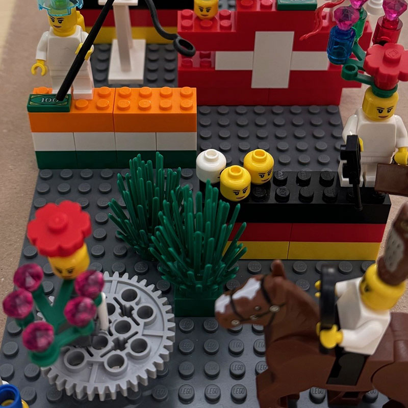 Detailreiche LEGO-Szene mit verschiedenen Mini-Figuren: Im Vordergrund eine Figur auf einem braunen Pferd, daneben andere Figuren mit Blumen auf dem Kopf. Mehrere Nationalflaggen aus LEGO-Steinen, darunter Deutschland, Indien und Schweiz, sind zu erkennen. Um die Figuren herum stehen Pflanzen, Zahnräder und lose LEGO-Köpfe