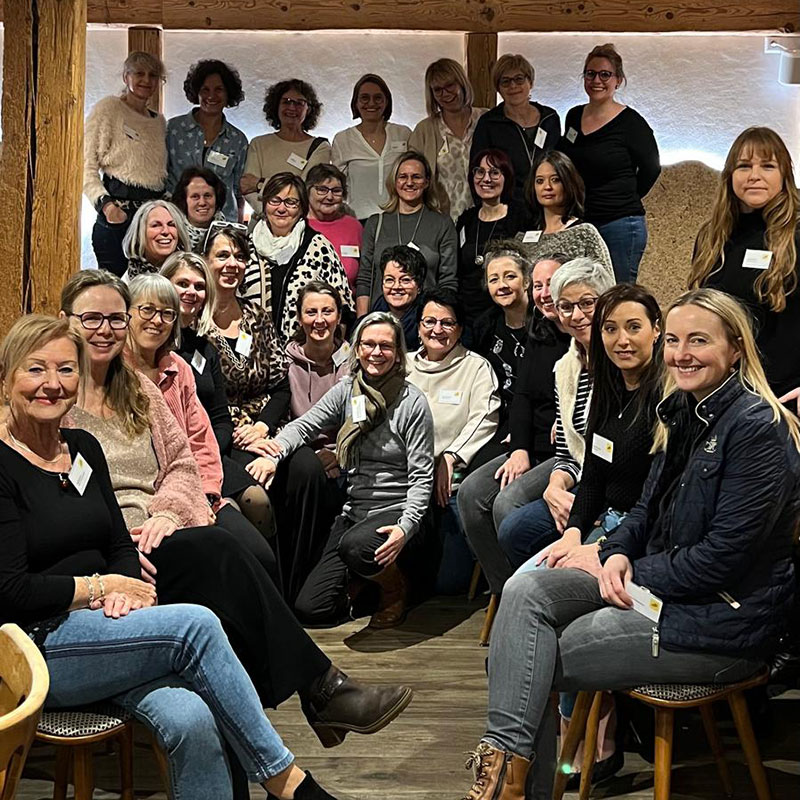 Eine große Gruppe von Frauen sitzt und steht dicht beisammen in einem gemütlich beleuchteten Raum mit Holzelementen. Alle lächeln in die Kamera und tragen Namensschilder, was auf eine Veranstaltung oder ein Seminar hindeutet. Die Atmosphäre wirkt freundlich und verbunden.