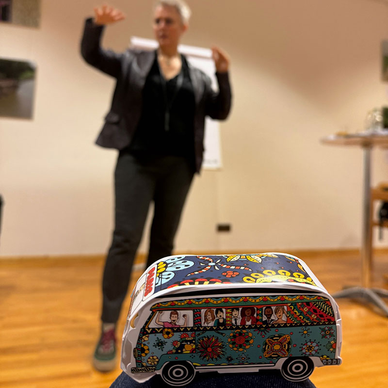 Im Vordergrund steht ein bunt bemalter, stilisierter Spielzeug-Bus mit Blumenmustern und Comic-Figuren auf den Fenstern. Im Hintergrund, unscharf dargestellt, steht eine Frau mit kurzen Haaren in einer dunklen Jacke und gestikuliert mit erhobenen Händen. Sie scheint vor einer Flipchart-Präsentation zu sprechen.