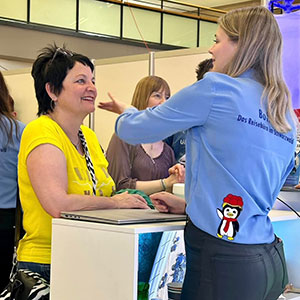 Eine Person in einem gelben Hemd spricht mit einer anderen Person in einem blauen Hemd, das den Text ‚Das Reiseland im Schwarzwald‘ und einen Pinguin zeigt. Szene an einem Kundenservice-Schalter bei einem Event.
