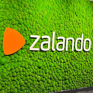 Moos-Wanddekoration mit dem weißen ‚Zalando‘-Logo in der Mitte und einem modernen Innenraum im Hintergrund.