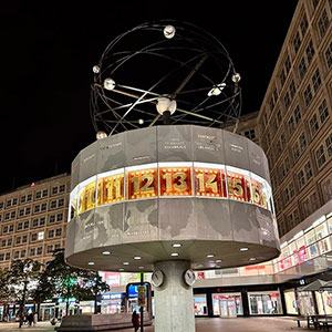 Die beleuchtete Weltzeituhr (Weltzeituhr) am Alexanderplatz in Berlin bei Nacht, umgeben von Gebäuden und Geschäften.