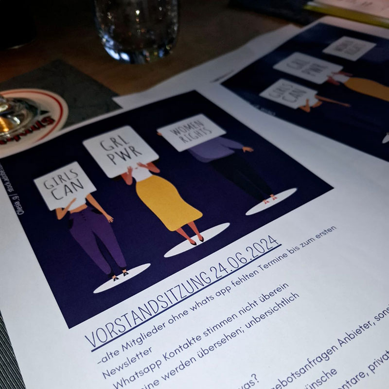 Gedrucktes Dokument mit der Überschrift ‚Vorstandssitzung 24.06.2024‘ auf einem Tisch, daneben ein Glas. Oben ist eine Illustration von drei Frauen mit Schildern, auf denen ‚Girls Can‘, ‚GRL PWR‘ und ‚Women Rights‘ steht. Darunter folgen Stichpunkte zu Themen der Sitzung.